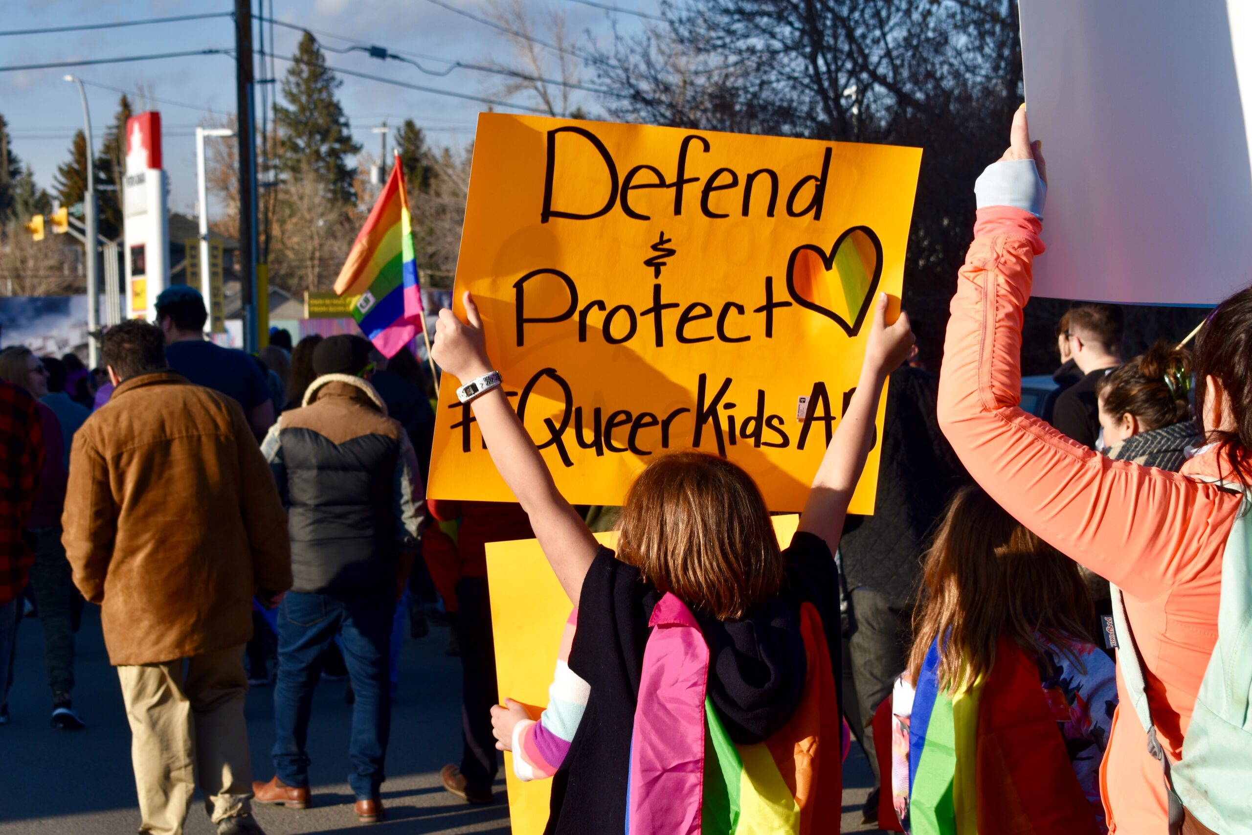 Image of people at a protest. One child holds up a sign that says Defend and Protect Queer Kids