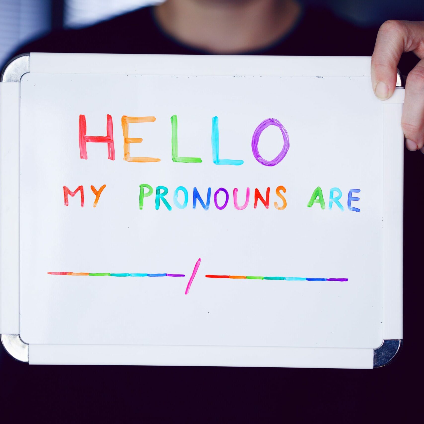 Image of a person holding a miniature size white board that says Hello my pronouns are blank/blank